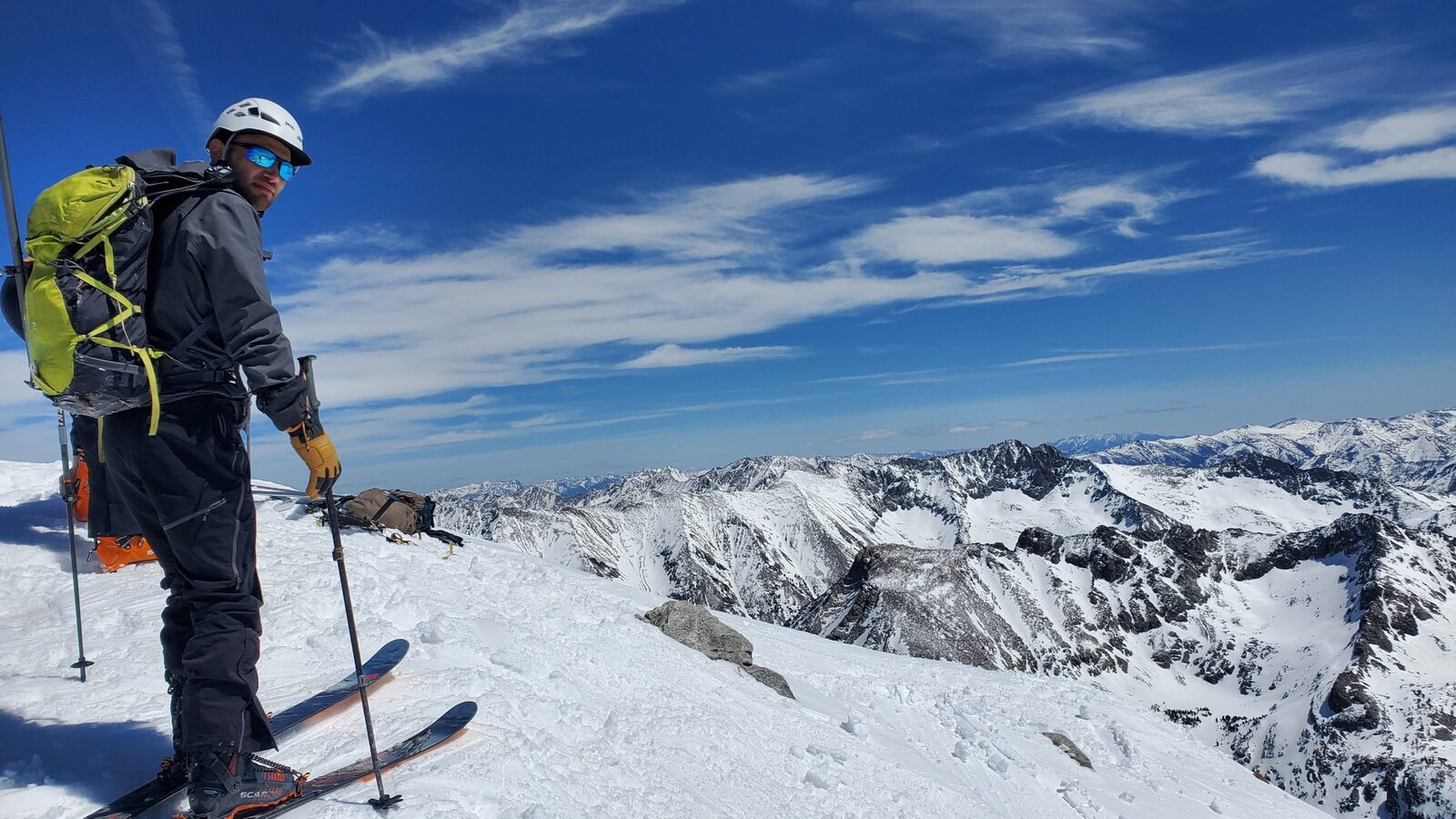 The maker of PowDash on a snowy summit with skis, peaks and blue sky behind